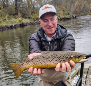An angler bundled up in a black winter jacket holding a beautiful Brown Trout caught while fly fishing on a cold day.